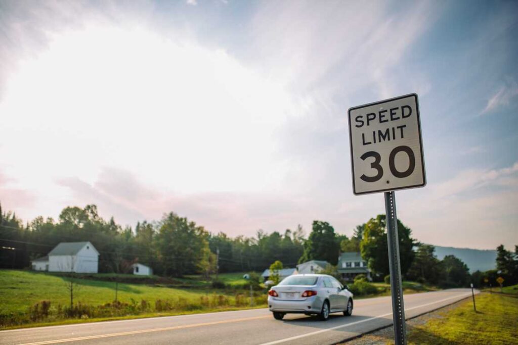 30 mph speed limit sign on country highway with small gray car driving past a farm with white buildings