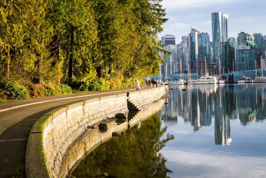 green trees and paved pathway along the water with boats and vancouver skyline in the background