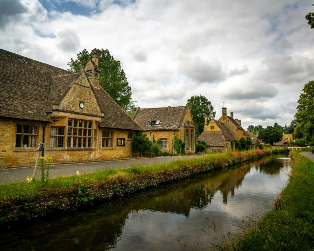 A brown and gray house beside a river under cloudy sky during daytime in Cotswolds. 
