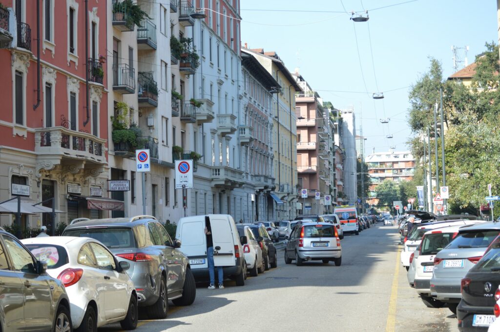A busy Milan street lined with parked cars next to tall, elegant buildings, showcasing the city’s vibrant urban life.