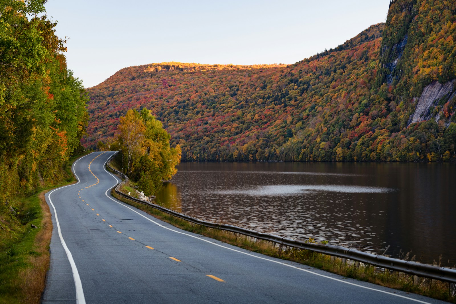 A winding road curves along a lake beneath hillsides of red, orange and yellow autumn foliage in New Hampshire.