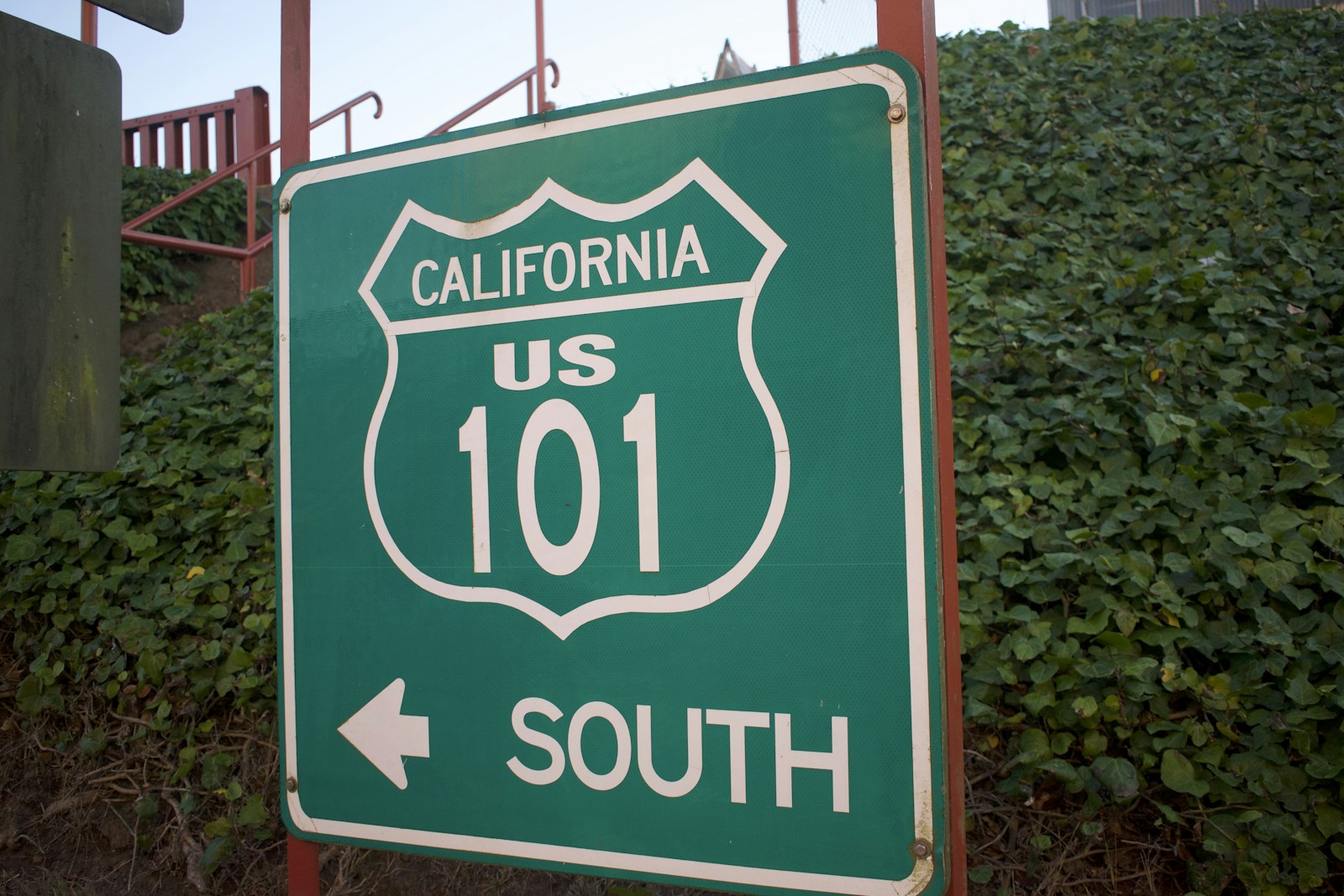 A green California US 101 South highway sign with an arrow pointing left, set against an ivy-covered wall